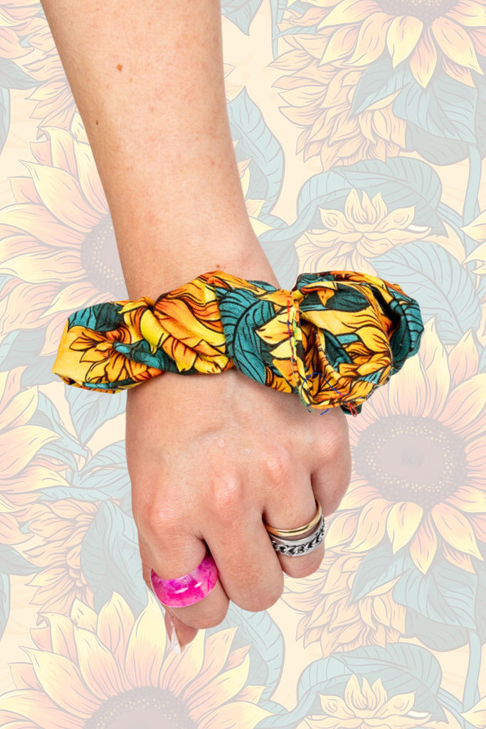 A yellow sunflower scrunchie with green leaves on model's wrist. The model is also wearing a pink and a silver ring. The background of the photo is the sunflower pattern.