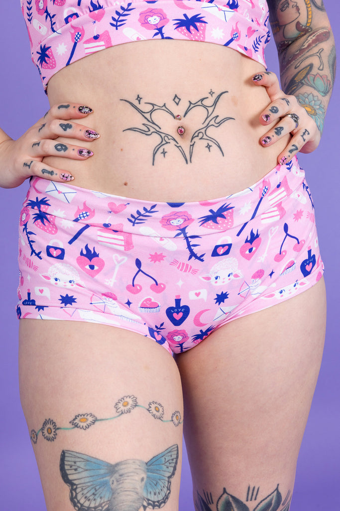 Close up of model wearing Wilde Mode x Amy Hastings Hipsters paired with matching comfort top. The fabric is baby pink with pink, white and purple kitsch tattoo designs, including lambs, hearts, cherries cake, strawberries and flowers. The background of the photo is purple. 
