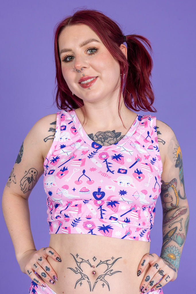 tattooed model with red hair wearing Wilde Mode x Amy Hastings Comfort Top. The fabric is baby pink with pink, white and purple kitsch tattoo designs, including lambs, hearts, cherries cake, strawberries and flowers. Model is smiling at the camera with her hands on her hips. The background of the photo is purple. 