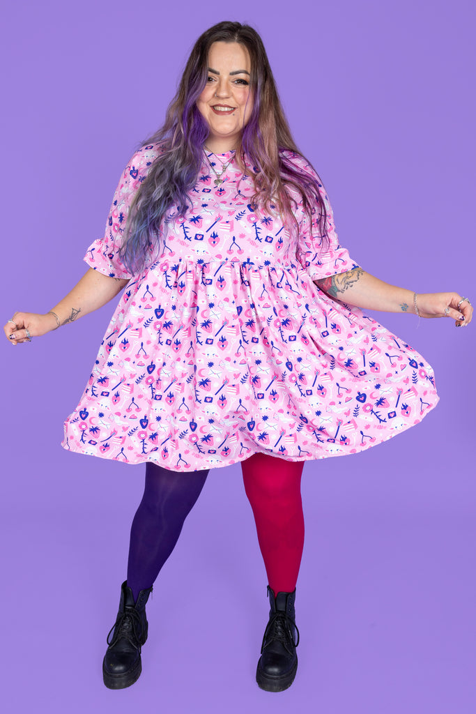 model with pastel purple hair is wearing Wilde Mode X Amy Hastings Dress paired with purple and pink tights and black boots. The fabric is baby pink with pink, white and purple kitsch tattoo designs, including lambs, hearts, cherries cake, strawberries and flowers. The model is smiling at the camera and swishing the dress. The background of the photo is purple. 