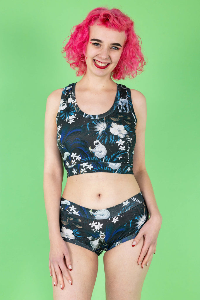 Lottie is a tall femme white pink haired model with a bob she is wearing  a matching crop top and briefs in a print on a navy base with cute monkeys and white flowers. The background is green and it is shot in a studio. She has her hands on her legs and is looking to camera