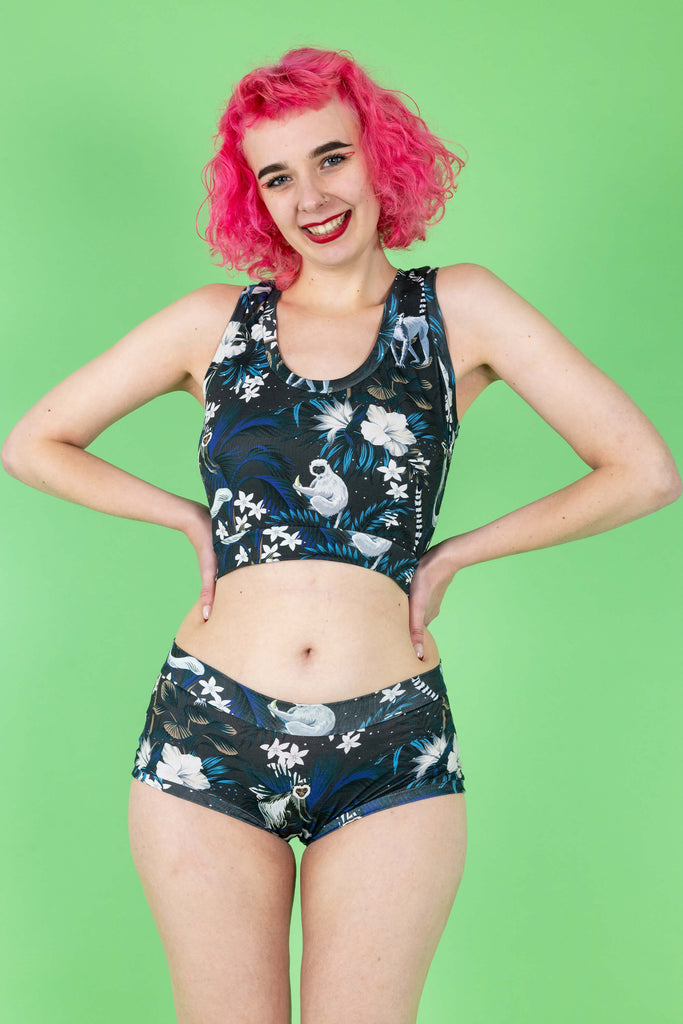 Lottie is a tall femme white pink haired model with a bob she is wearing  a matching crop top and hipster briefs in a print on a navy base with cute monkeys and white flowers. The background is green and it is shot in a studio. She has her hands on her hips and is looking to camera