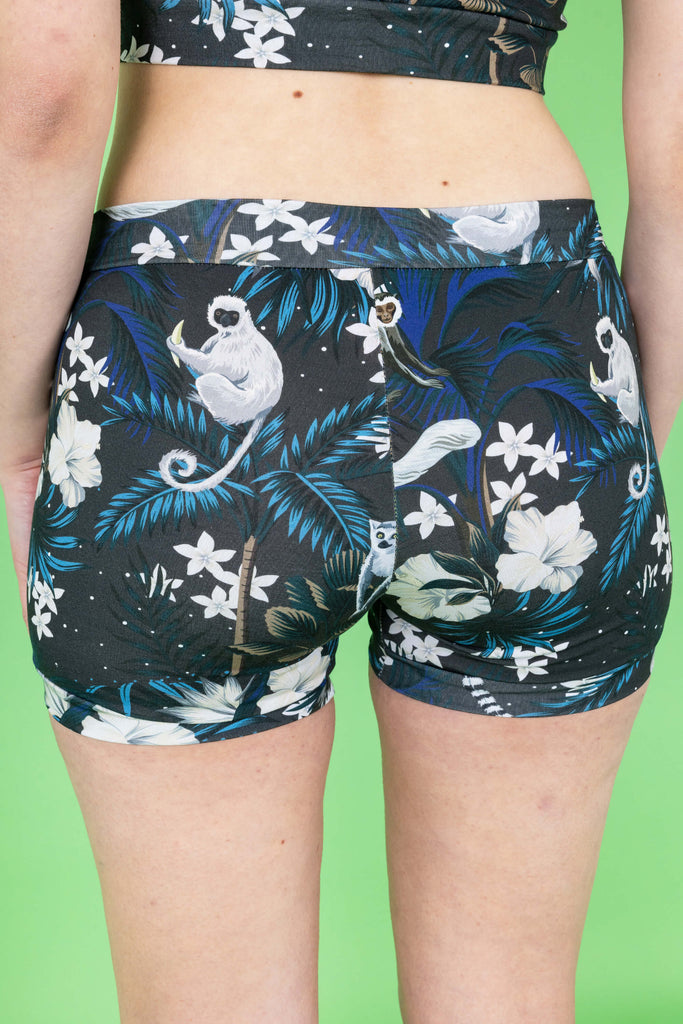 Lottie is a tall femme white pink haired model with a bob she is wearing  a matching crop top and boxers in a print on a navy base with cute monkeys and white flowers. The background is green and it is shot in a studio.This is a close up of her bottom in the boxers