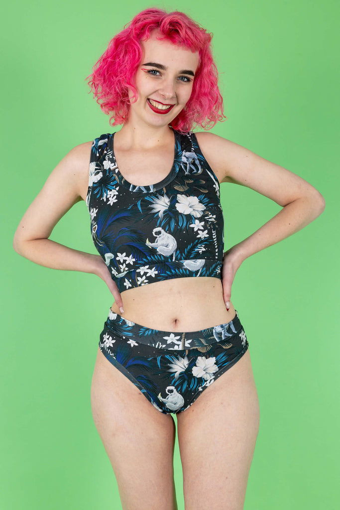 Lottie is a tall femme white pink haired model with a bob she is wearing  a matching crop top and thong in a print on a navy base with cute monkeys and white flowers. The background is green and it is shot in a studio. She has her hands on her hips and is looking to camera