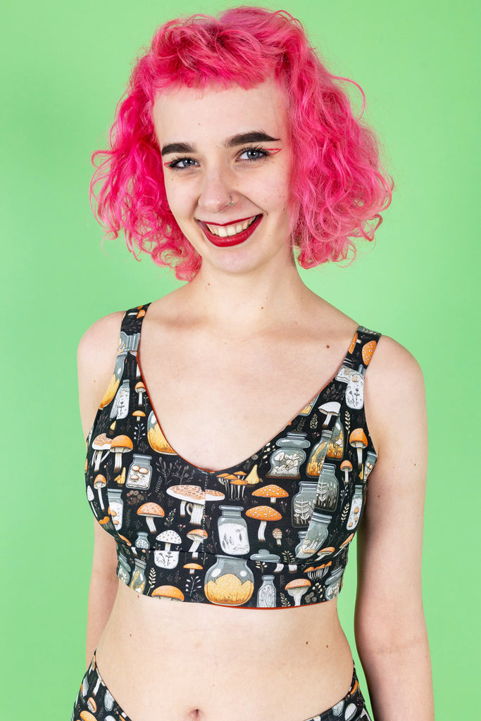 Lottie a tall white pink haired model is wearing a mushroom potions print on a black background two piece underwear set smiling at camera. The background is green.