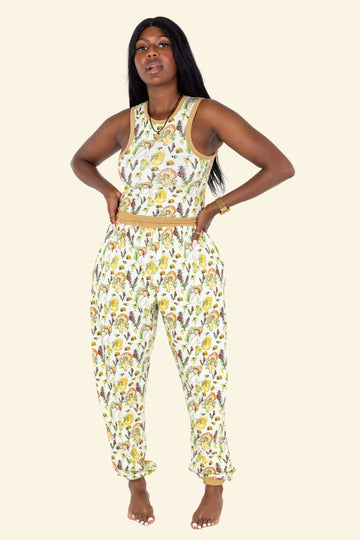 Model with long black hair wearing Wilde Mode x The Mushroom Babes Joggers. The print on the joggers is of various mushroom babes surrounded by flowers and insects. The banding is a mustard yellow. The model is posing with her hands on her hips.
