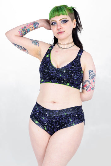 Tattooed model with green hair wearing Purple Spider Comfort Lounge Bra. The print has a black background with purple spiderwebs, green spiders and white starts all over. Model is posing with hand in hair and other hand behind back.