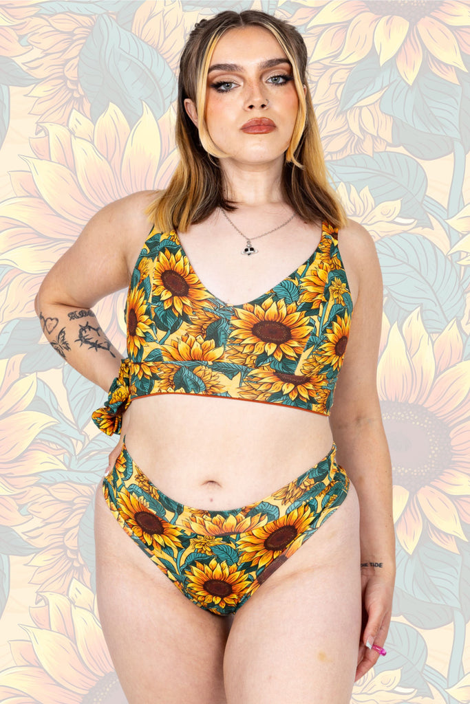 Model is wearing Sunflower Field Comfort Bra with matching thong. The print is a yellow background with big yellow sunflowers with a brown centre and green leaves. Model is posing facing the camera with one hand on their hip. The background of the photo is a faded close up of the Sunflower Field print.