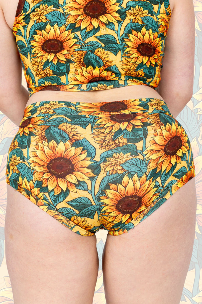 Close up of Sunflower Field Comfort Hipsters shown on a model standing with their back to the camera to show back of hipsters. The print is a yellow background with big yellow sunflowers with a brown centre and green leaves. The background of the photo is a faded close up of the Sunflower Field print.