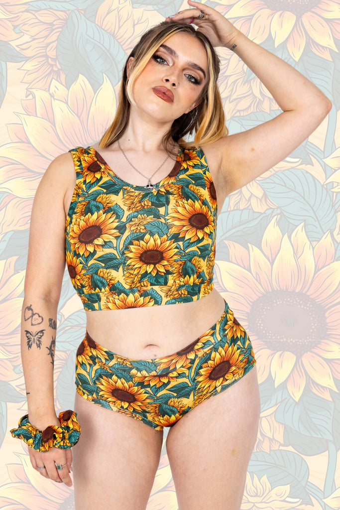 Model wearing Sunflower Field Comfort top paired with matching hipsters and scrunchie. The print is a yellow background with big yellow sunflowers with a brown centre and green leaves. The model is posing with one. The background of the photo is a faded close up of the Sunflower Field print.