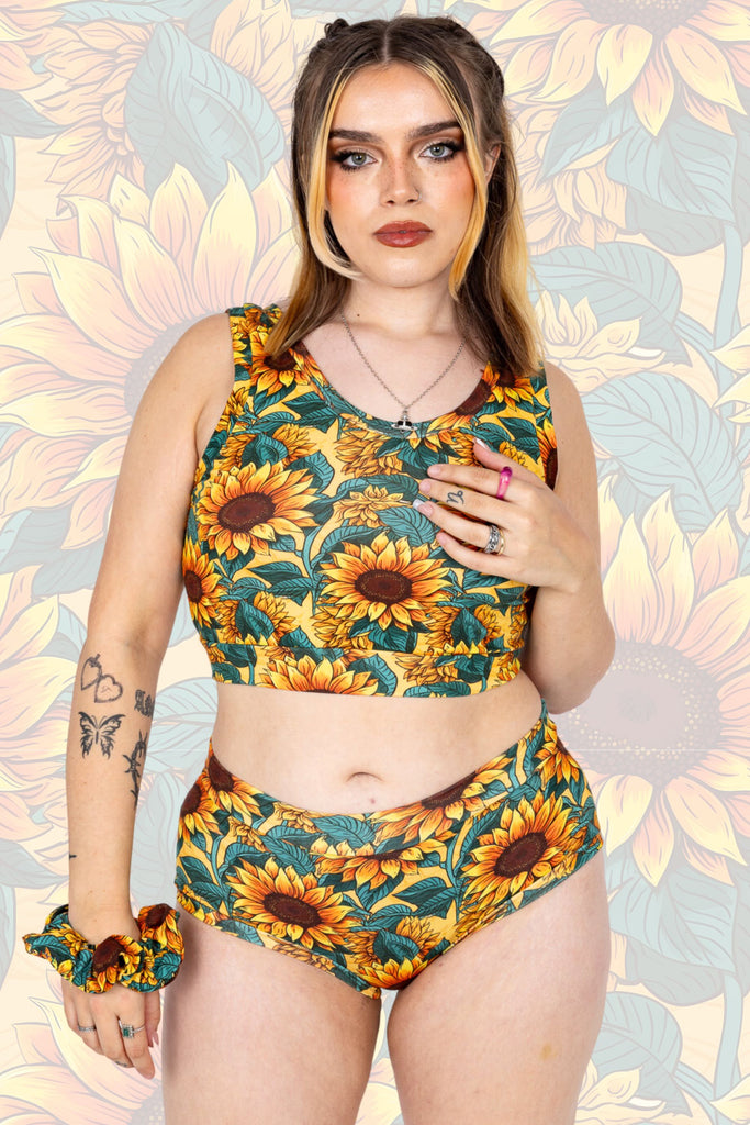 Model wearing Sunflower Field Comfort top paired with matching hipsters and scrunchie. The print is a yellow background with big yellow sunflowers with a brown centre and green leaves. The model is posing with one hand by their side and other resting on the comfort top. The background of the photo is a faded close up of the Sunflower Field print.