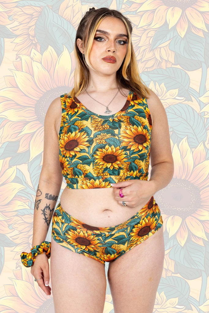 Tattooed model is wearing Sunflower Field Comfort Briefs paired with matching comfort top and scrunchie. The print is a yellow background with big yellow sunflowers with a brown centre and green leaves. The model is posing with one hand by their side and other playing with top. The background of the photo is a faded close up of the Sunflower Field print.   