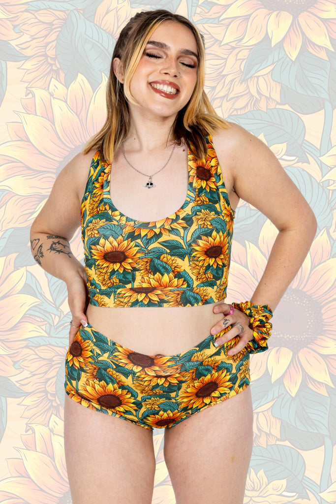 Model wearing Sunflower Field Comfort Briefs paired with matching comfort top and scrunchie. The print is a yellow background with big yellow sunflowers with a brown centre and green leaves. The model is smiling with eyes closed and hands on hips.  The background of the photo is a faded close up of the Sunflower Field print.