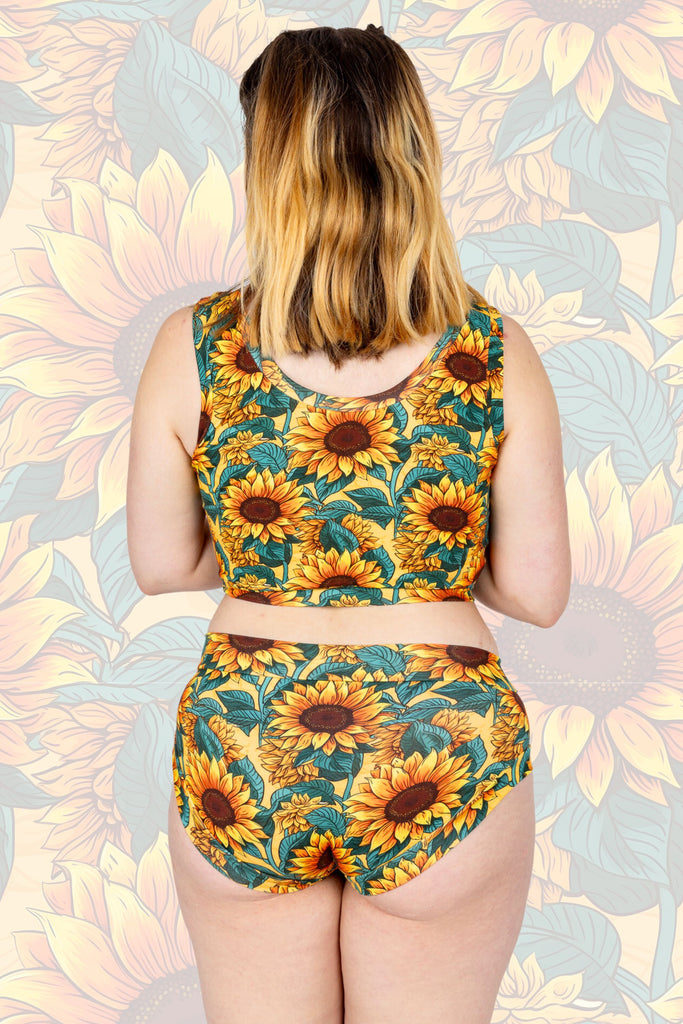 Model is wearing Sunflower Field Comfort Hipsters paired with matching comfort top. The print is a yellow background with big yellow sunflowers with a brown centre and green leaves. Model is standing with their back to the camera to show the back of the hipsters. The background of the photo is a faded close up of the Sunflower Field print.