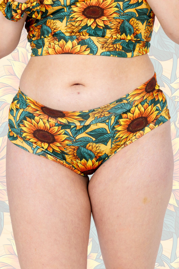 Close up of Sunflower Field Comfort Briefs shown on a model facing the camera. The print is a yellow background with big yellow sunflowers with a brown centre and green leaves. The background of the photo is a faded close up of the Sunflower Field print.