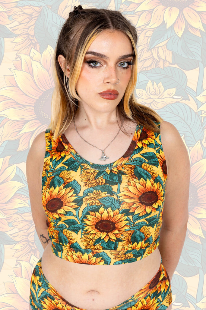 Close up of model wearing Sunflower Field Comfort Top whilst  facing the camera. The print is a yellow background with big yellow sunflowers with a brown centre and green leaves. The model is posing with one hand by their side and other resting on the comfort top. The background of the photo is a faded close up of the Sunflower Field print.