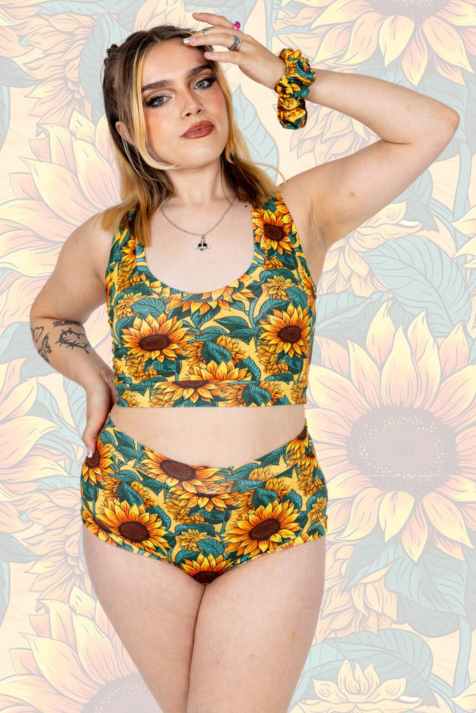 Model with blonde hair is wearing Sunflower Field Comfort Briefs paired with matching comfort top and scrunchie. The print is a yellow background with big yellow sunflowers with a brown centre and green leaves. Model is posing with one hand on their hip and other in their hair. Model is looking at the camera. The background of the photo is a faded close up of the Sunflower Field print.