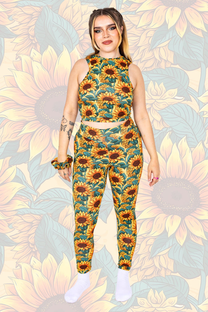 Model is wearing Sunflower Field Leggings paired with matching racer front top and scrunchie on their wrist. The print is a yellow background with big yellow sunflowers with a brown centre and green leaves. Model is standing with hands by their sides and smiling at the camera. The background of the photo is a faded close up of the Sunflower Field print.