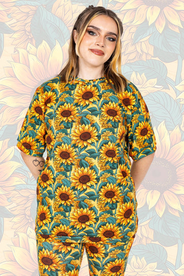 Tattooed model wearing Sunflower Field T-Shirt paired with matching leggings. The print is a yellow background with big yellow sunflowers with a brown centre and green leaves. The model is smiling at the camera with their hands behind their back.