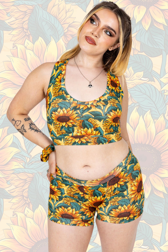 Tattooed model with cool makeup and blonde hair wearing Sunflower Field Boxers paired with matching comfort top and scrunchie. The print is a yellow background with big yellow sunflowers with a brown centre and green leaves. The model is posing with one hand on their hip and looking into the distance.