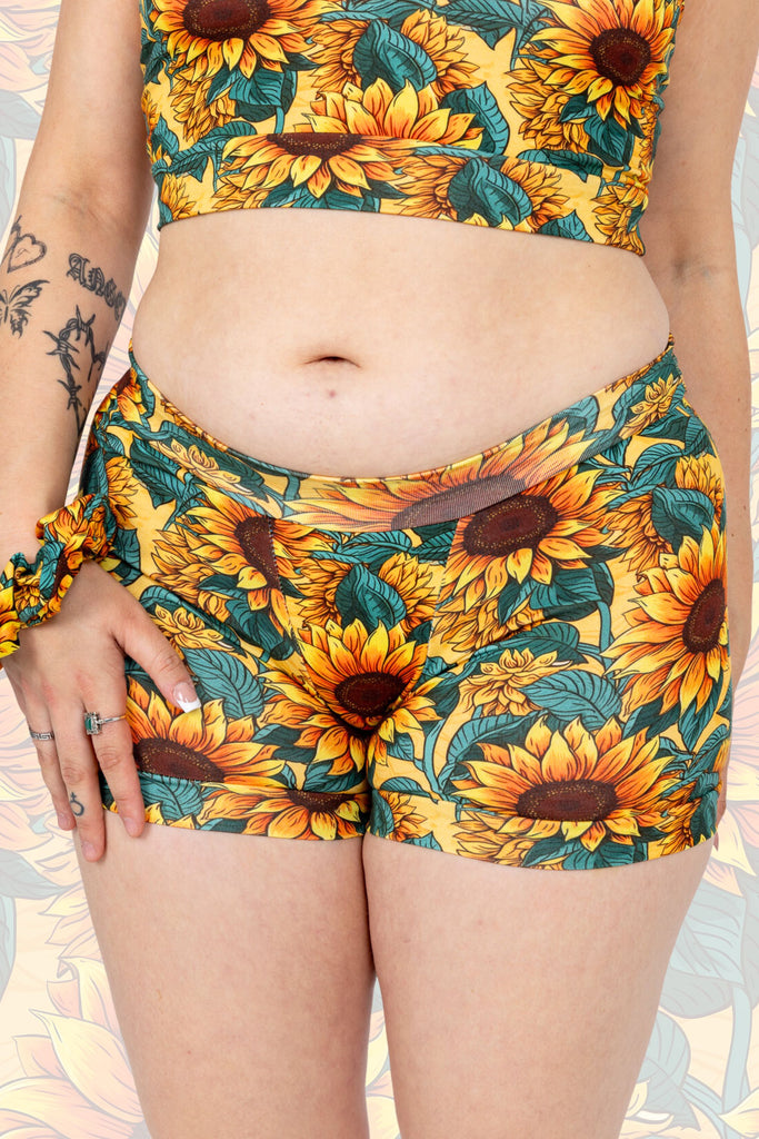 Close up of model wearing Sunflower Field Boxers. The print is a yellow background with big yellow sunflowers with a brown centre and green leaves. Model is posing facing the camera with matching scrunchie on wrist which is resting on their thigh.