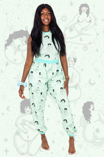 Model is wearing Wilde Mode x Fat Lemon Mint Celestial Beauties Joggers paired with matching racer front top and scrunchie on their wrist. The print is of various naked people amongst stars with mint green banding. Model is posing and smiling at camera. The background of the photo is a close up of the print.