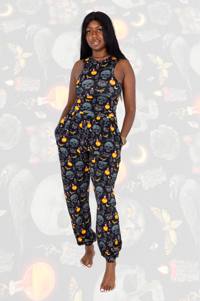 Model is wearing Black Magic Joggers paired with matching racer front top. The print is a black background with all over pattern of skulls, black cats, orange potion bottles, crescent moons, moths and candles. Model is posing with hands in pockets of the joggers and one leg in front of the other. The background of the photo is a faded close up of the print.