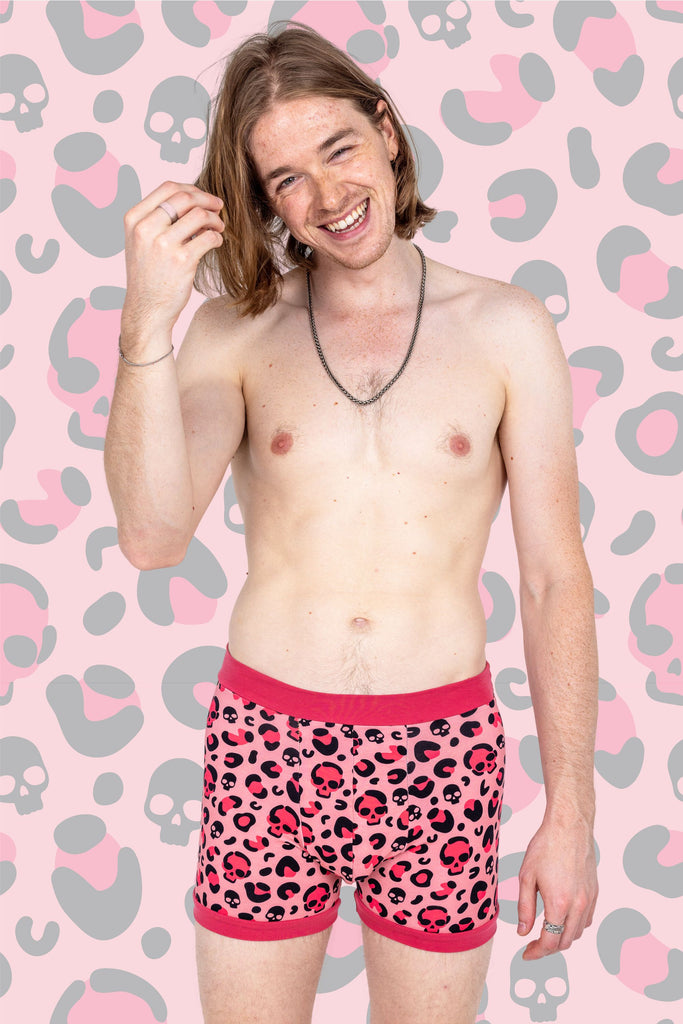 Model wearing Punky Babe Leopard Print Boxers, the boxers are a baby pink colour with all over hot pink and black leopard print and skull print with hot pink bands. The background of the photo is a close up of the print. Model is smiling with hand in his hair.