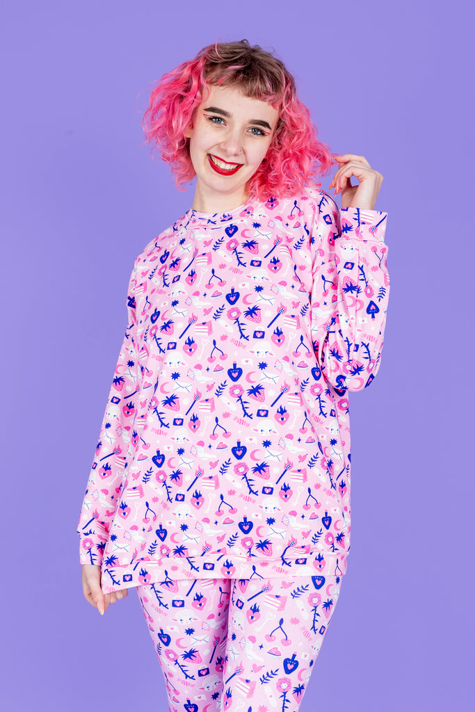 Model with pink hair and fun makeup is wearing Wilde Mode x Amy Hastings Lounge Jumper paired with matching leggings. The fabric is baby pink with pink, white and purple kitsch tattoo designs, including lambs, hearts, cherries cake, strawberries and flowers. The model is smiling at the camera with her hand in her hair. The background of the photo is purple.