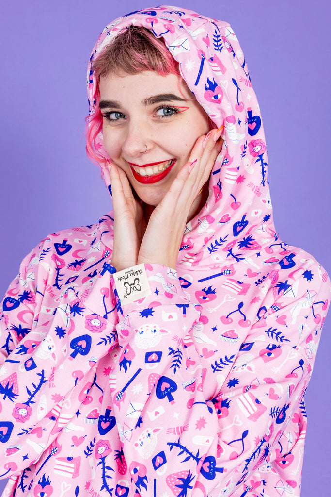 Close up of model with pink hair and fun makeup wearing Wilde Mode x Amy Hastings Hoodie. The fabric is baby pink with pink, white and purple kitsch tattoo designs, including lambs, hearts, cherries cake, strawberries and flowers. The close up is of the model looking cosy with her hood up and smiling with her hands on her cheeks, looking at the camera. The background of the photo is purple. 