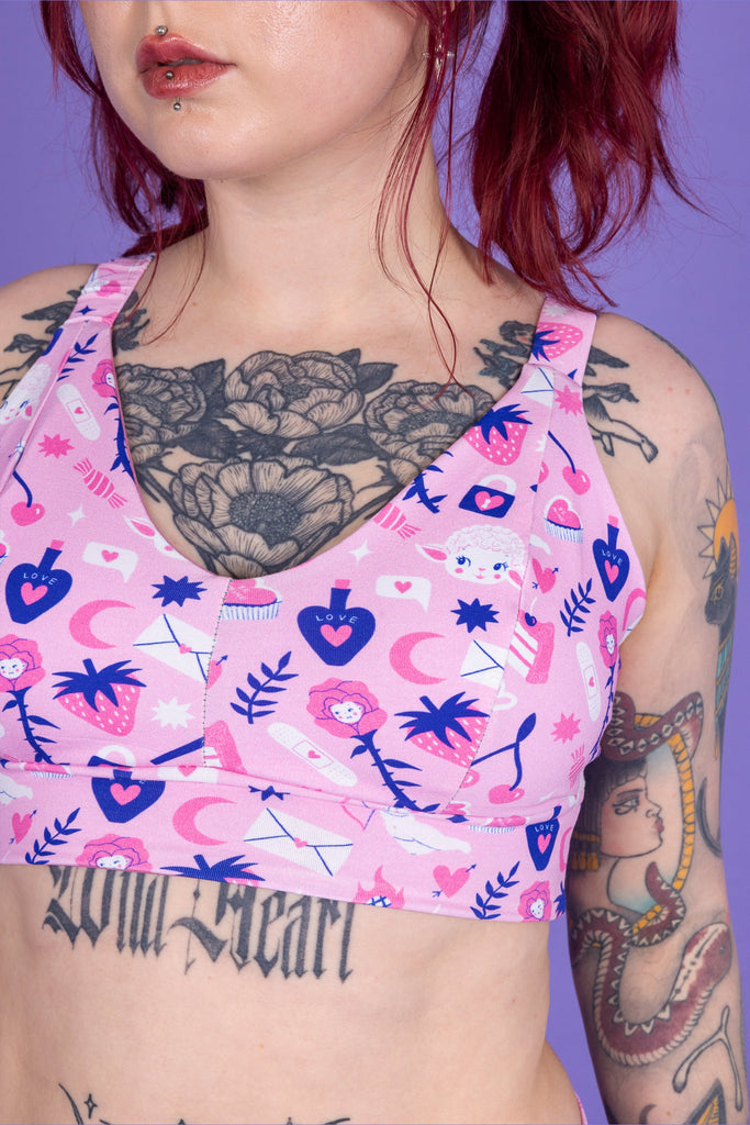 Close up of tattooed model with red hair wearing Wilde Mode x Amy Hastings Comfort Bra. The fabric is baby pink with pink, white and purple kitsch tattoo designs, including lambs, hearts, cherries cake, strawberries and flowers. 