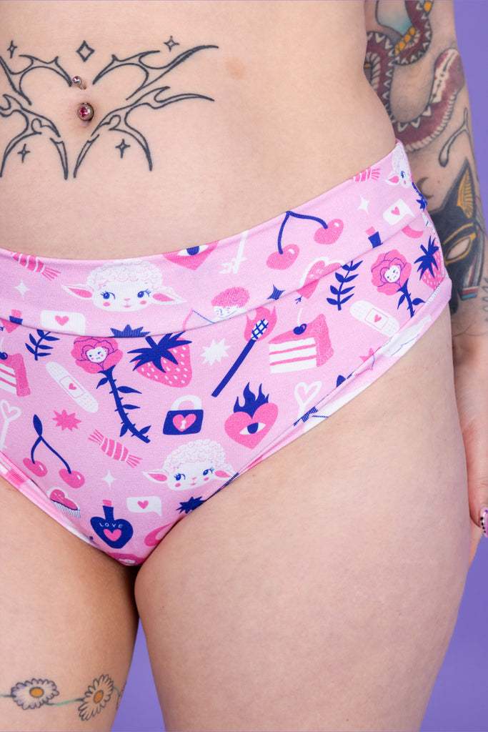 Close up of tattooed model with red hair wearing Wilde Mode x Amy Hastings Comfort Thong. The fabric is baby pink with pink, white and purple kitsch tattoo designs, including lambs, hearts, cherries cake, strawberries and flowers. Close up shows the front of the thong in more detail. 