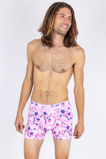 male model with long hair wearing Wilde Mode x Amy Hastings Boxers. The fabric is baby pink with pink, white and purple kitsch tattoo designs, including lambs, hearts, cherries cake, strawberries and flowers. The model is smiling and looking into the distance. The background of the photo is white. 