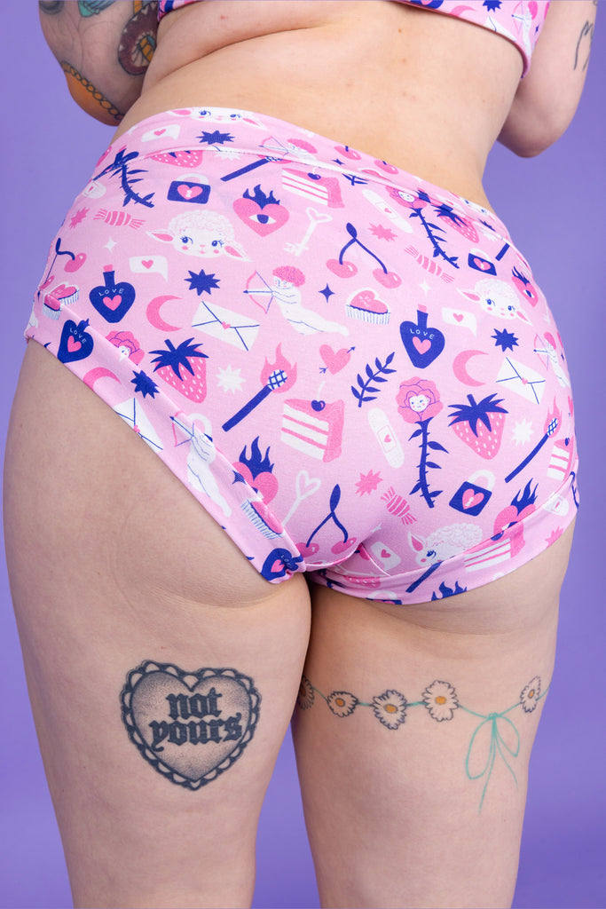 Close up of tattooed model wearing Wilde Mode x Amy Hastings Comfort Briefs. The fabric is baby pink with pink, white and purple kitsch tattoo designs, including lambs, hearts, cherries cake, strawberries and flowers. Close up shows model with their back to the camera to show the back of the briefs. 