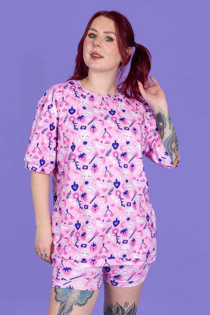 tattooed model with red hair is wearing Wilde Mode x Amy Hastings Tee paired with matching boxers. The fabric is baby pink with pink, white and purple kitsch tattoo designs, including lambs, hearts, cherries cake, strawberries and flowers. The model is posing with one hand in her hair and is smiling at the camera. The background of the photo is purple. 
