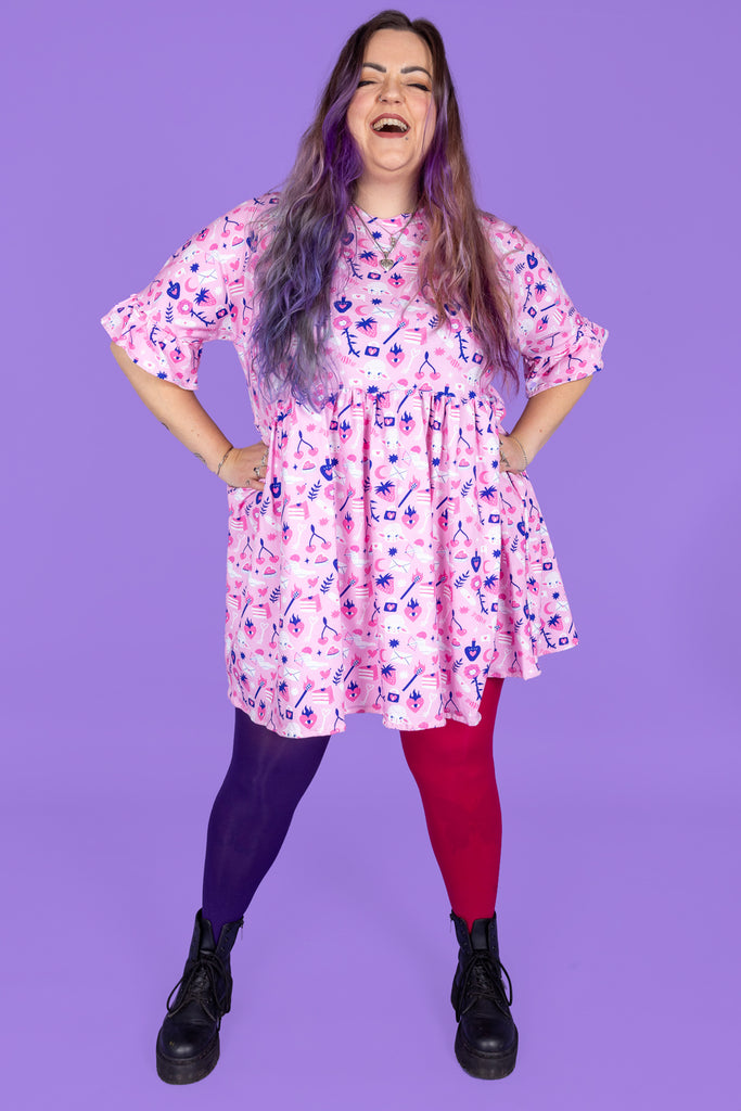 model with pastel purple hair is wearing Wilde Mode X Amy Hastings Dress paired with purple and pink tights and black boots. The fabric is baby pink with pink, white and purple kitsch tattoo designs, including lambs, hearts, cherries cake, strawberries and flowers. Model is laughing with her hands on her hips. The background of the photo is purple. 