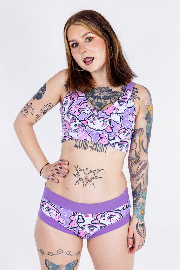 Tattooed model wearing Wilde Mode x Twinkle and Gloom Lilac Angry Feminist Comfort Lounge Bra and Brief. The print is of angry pink cartoon faces holding up middle fingers on a purple background. The band on the brief is also purple.