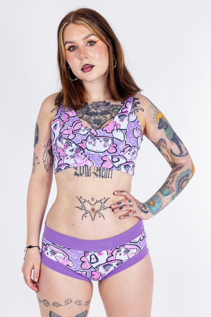 Tattooed model wearing Wilde Mode x Twinkle and Gloom Lilac Angry Feminist Comfort Lounge Bra and Brief. The print is of angry pink cartoon faces holding up middle fingers on a purple background. The band on the brief is also purple.