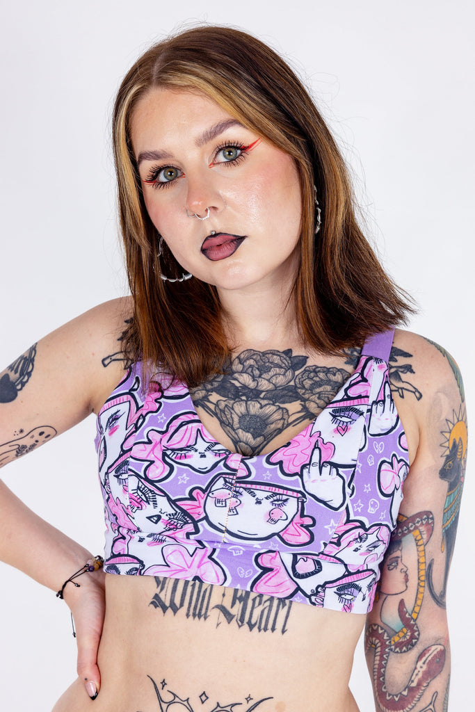 Tattooed model wearing Wilde Mode x Twinkle and Gloom Lilac Angry Feminist Comfort Lounge Bra. The print is of angry pink cartoon faces holding up middle fingers on a purple background. 