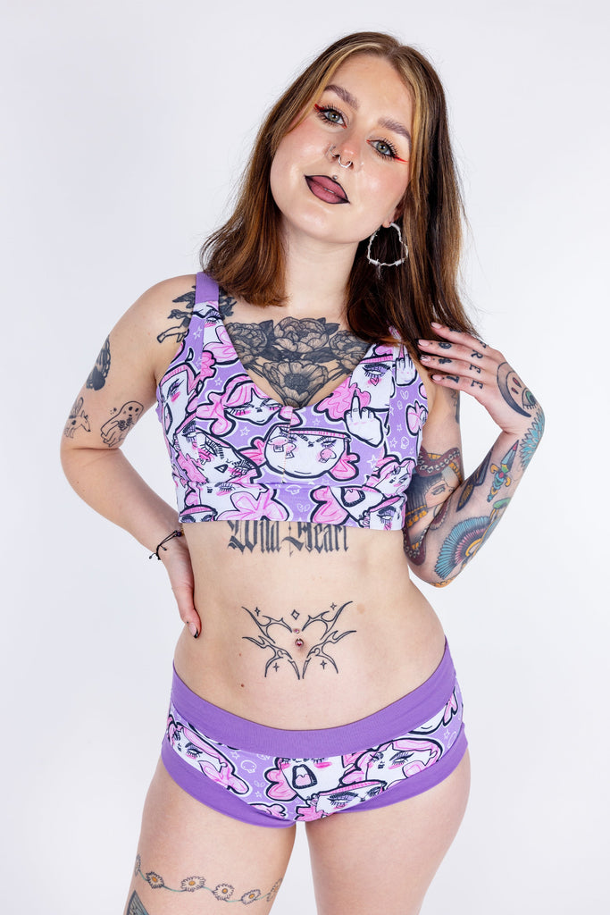 Tattooed model wearing Wilde Mode x Twinkle and Gloom Lilac Angry Feminist Comfort Lounge Bra and Brief. The print is of angry pink cartoon faces holding up middle fingers on a purple background. The band on the brief is also purple.