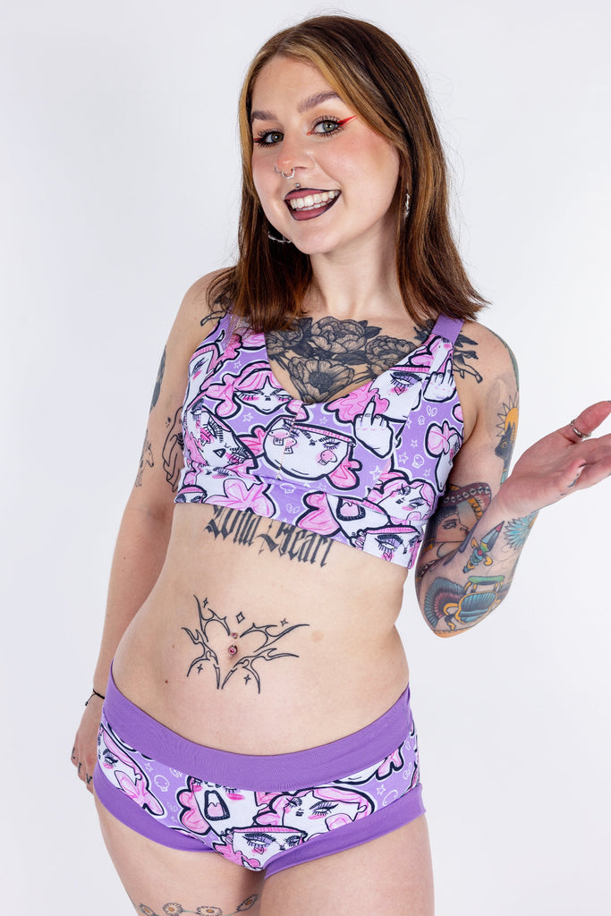 Tattooed model wearing Wilde Mode x Twinkle and Gloom Lilac Angry Feminist Comfort Lounge Bra and Brief. The print is of angry pink cartoon faces holding up middle fingers on a purple background. The band on the brief is also purple.