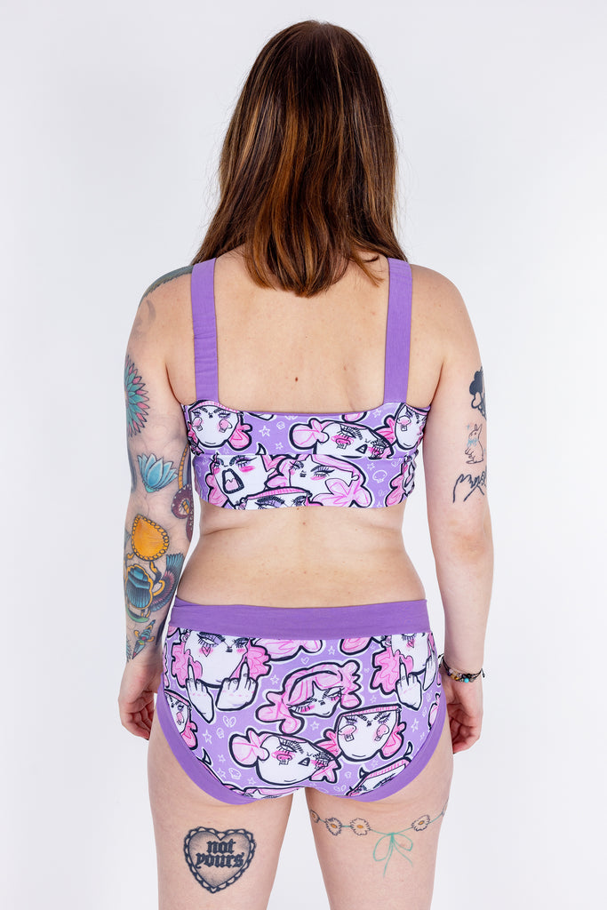 Tattooed model wearing Wilde Mode x Twinkle and Gloom Lilac Angry Feminist Comfort Briefs and Bra. The print is of angry pink cartoon faces holding up middle fingers on a purple background. The band on the brief is also purple.