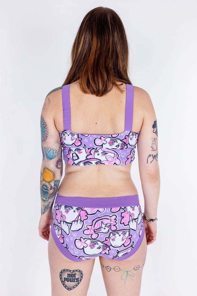 Tattooed model wearing Wilde Mode x Twinkle and Gloom Lilac Angry Feminist Comfort Lounge Bra and Brief. The print is of angry pink cartoon faces holding up middle fingers on a purple background. The band on the brief is also purple.