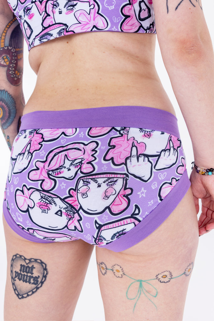 Tattooed model wearing Wilde Mode x Twinkle and Gloom Lilac Angry Feminist Comfort Briefs. The print is of angry pink cartoon faces holding up middle fingers on a purple background. The band on the brief is also purple.