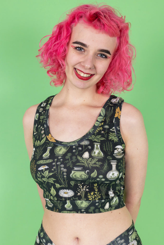 A close up of a model with pink hair, red eyeliner and red lipstick wearing the wilde mode apothecary comfort top with the matching boxers. The print features various green and yellow plants in vases on a dark green background. They are facing forward with both hands resting behind their back and smiling to camera.