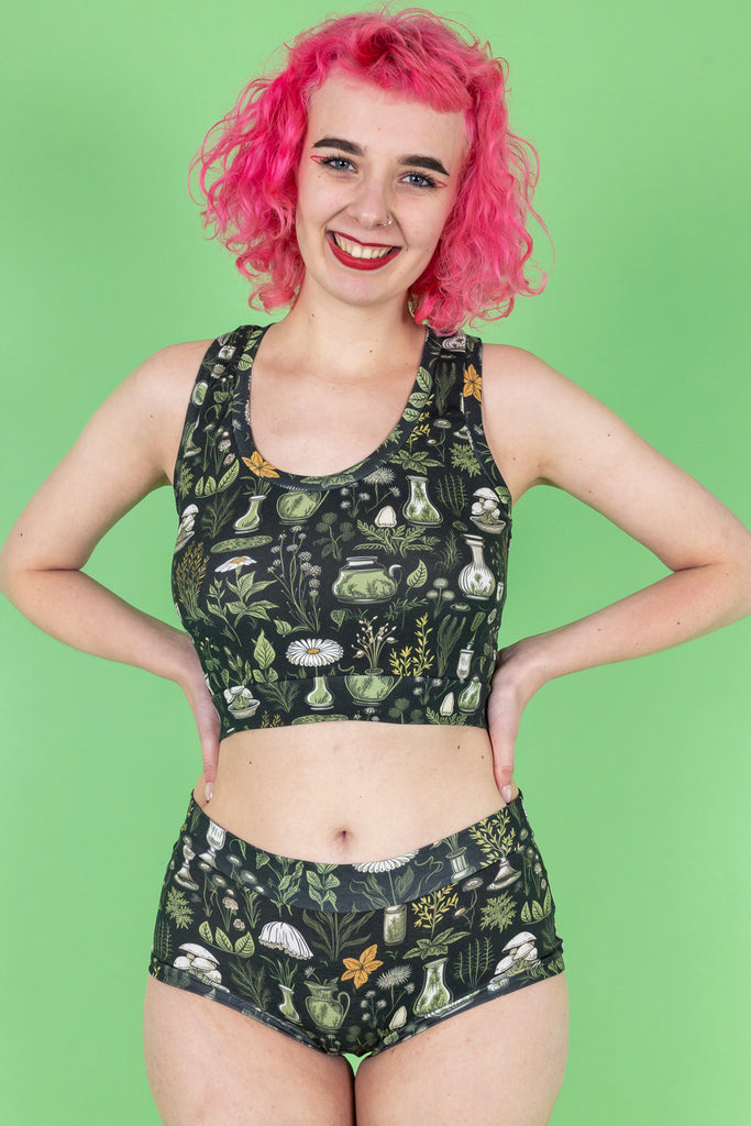 A model with pink hair, red eyeliner and red lipstick wearing the wilde mode apothecary comfort hipsters with the matching comfort top. The print features various green and yellow plants in vases on a dark green background. They are facing forward with both hands resting on their hips and smiling to camera.