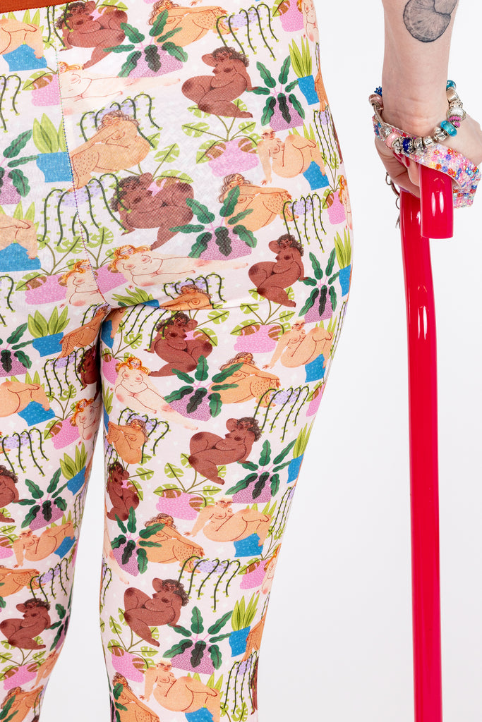 Closeup of Tattooed model disabled Eliza with rainbow coloured hair wearing Wilde Mode x Bunny Weller Leggings and comfort top. The print is of various naked ladies with different skin tones amongst various different plants. The waist and leg bands are a warm brown colour.
