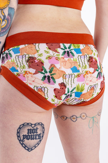 Tattooed model wearing Wilde Mode x Bunny Weller Comfort Briefs. The print is of various naked ladies with different skin tones amongst various different plants. The waist and leg bands are a warm brown colour.