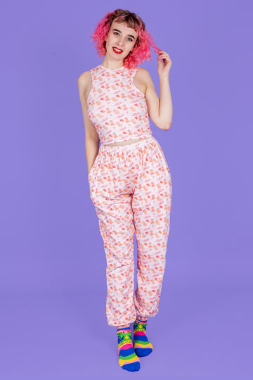 model with pink hair wearing Dinos in Love Joggers paired with matching racer front top. The fabric is white and the design is of little orange, pink and muted red dinosaurs with red and orange love hearts and speckles dotted around. The model is posing with one hand in her hair and the other hand in her pocket. he background of the photo is purple. 