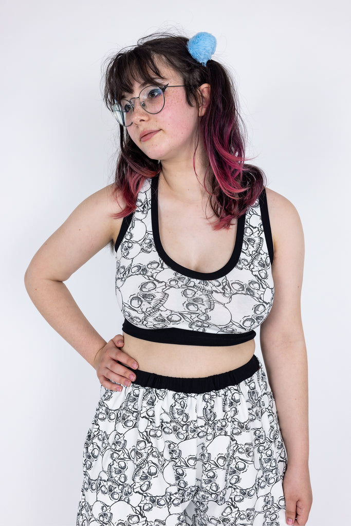 White Skulls of the Dead comfort crop top, with black skull print pattern and black hem. Worn as matching loungewear pyjama set with jogger bottoms, by model with brown and pink hair in bunches and heavy eyeliner wearing glasses, posing with hand on hip. Made sustainably in Scotland UK by Wilde Mode